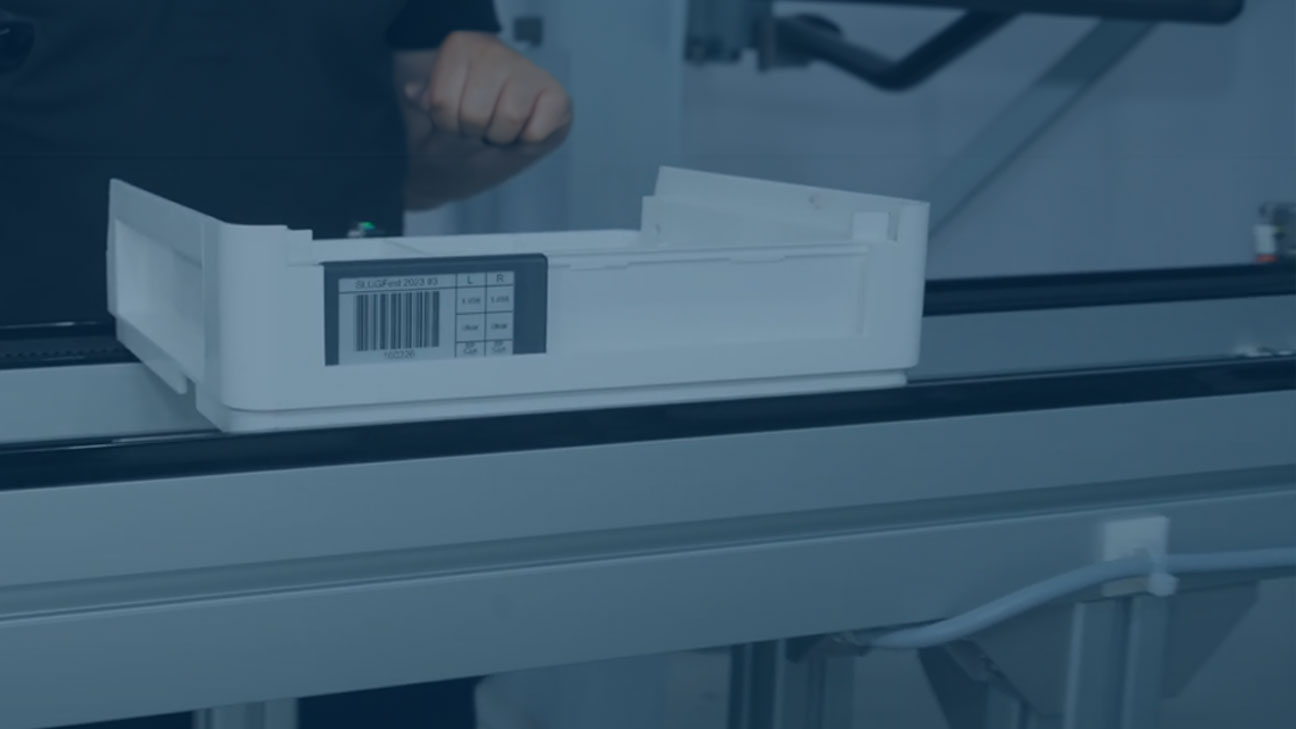 White container with a barcode label placed on a conveyor belt in a laboratory or industrial setting.